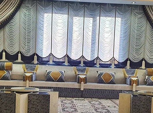 Cream-backed majlis with grey-blue bases and pillows below white festoon blinds, with a patterned rug and glass-topped square tables