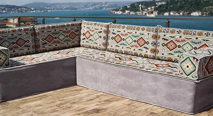Grey L-shaped floor sofa on a tall velvet base with kilim-weave seats, backrests and square armrest bolsters on a sunny terrace