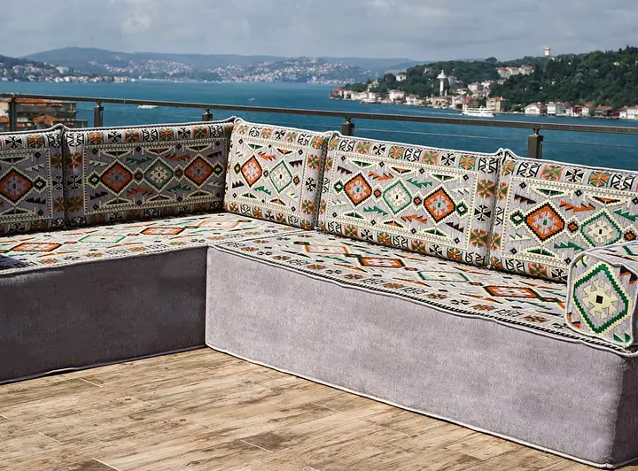 Grey L-shaped floor sofa on a tall velvet base with kilim-weave seats, backrests and square armrest bolsters on a sunny terrace