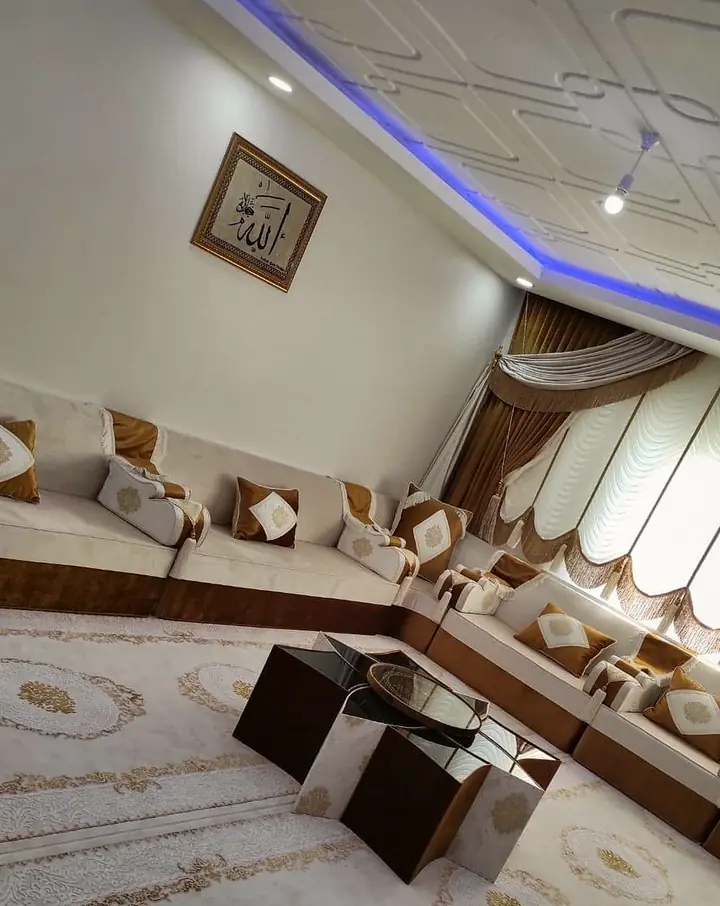 Corner of an ivory and brown majlis with gold diamond pillows, black cube tables and framed calligraphy beneath a blue cove light