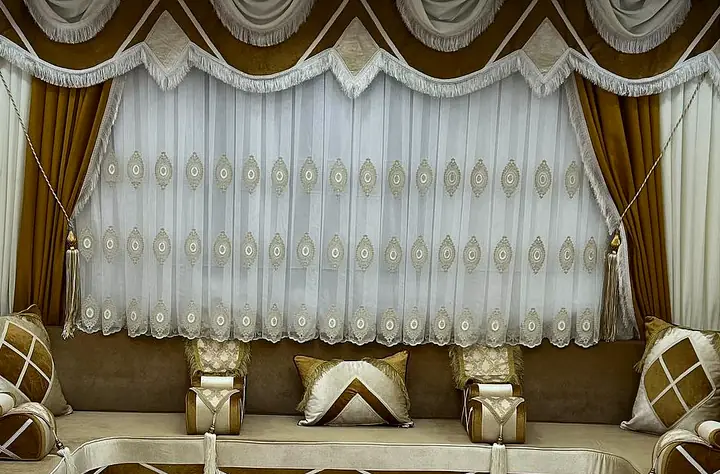 Ivory and caramel majlis run with white X-line pillows and cylinder armrests below embroidered sheer curtains and caramel drapes