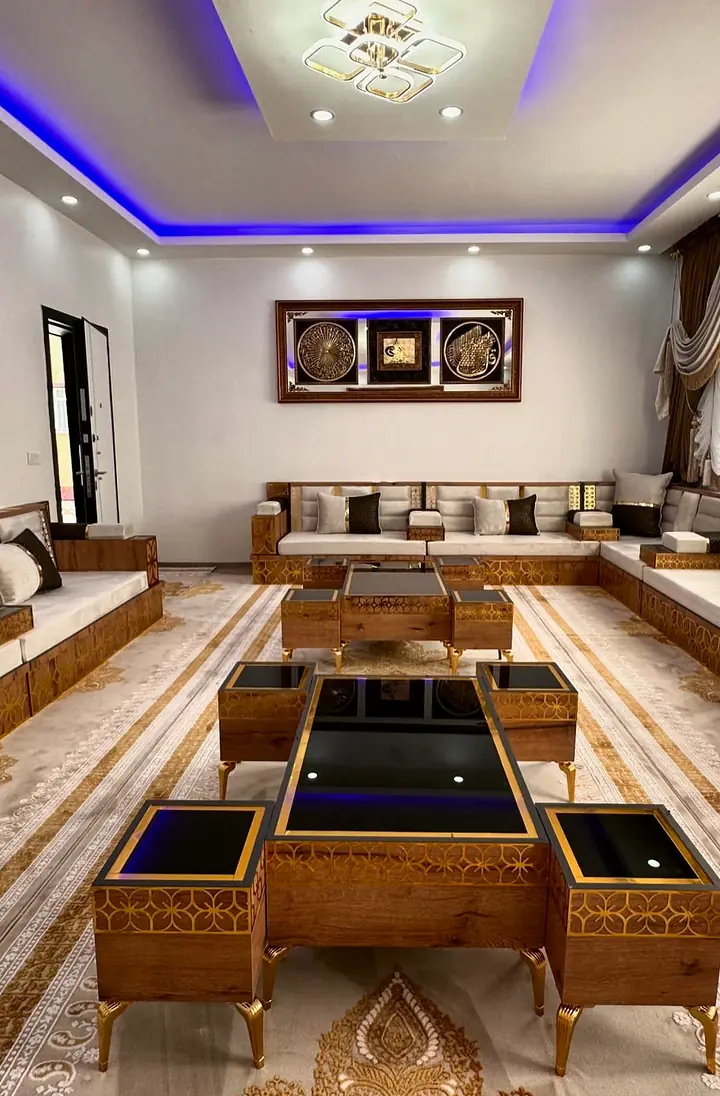 Long ivory majlis running down both walls of a hall with a blue lit ceiling cove and black glass nesting tables in the center