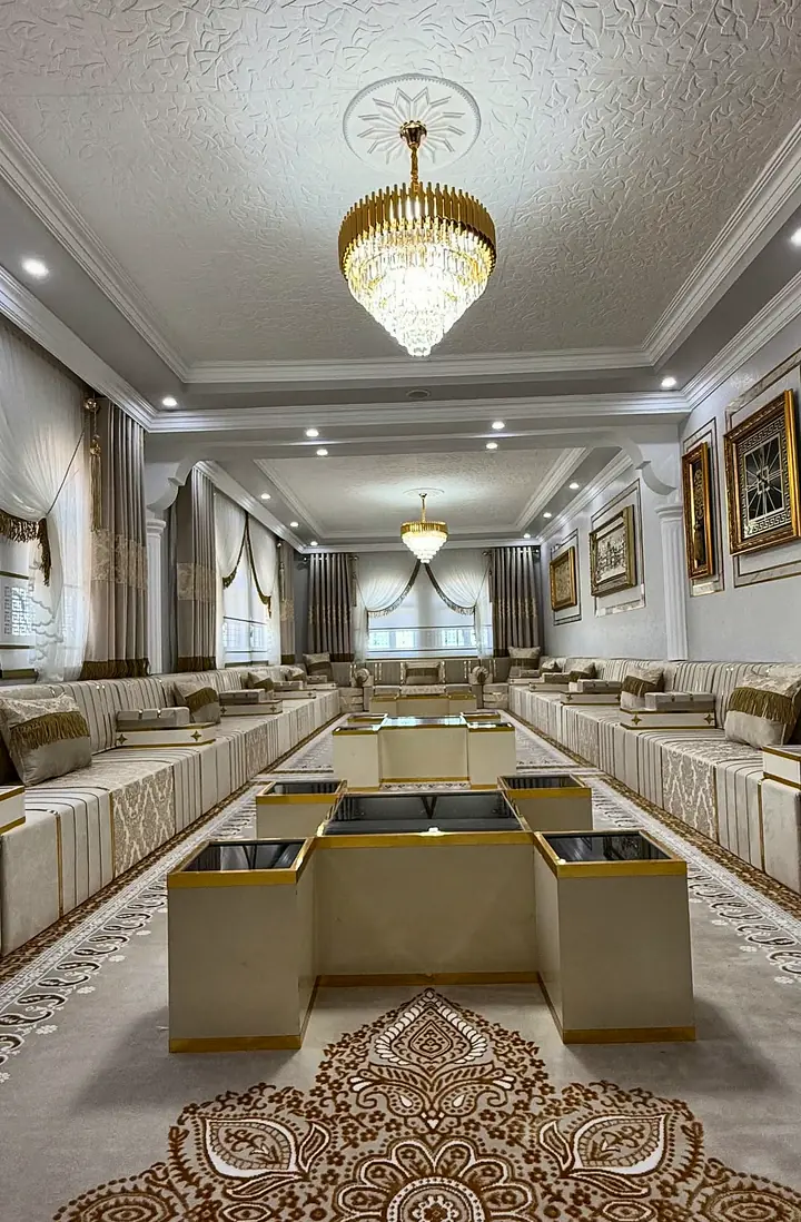 Long ivory damask majlis hall under crystal chandeliers, with cream cube tables topped in black glass and a large gold medallion rug