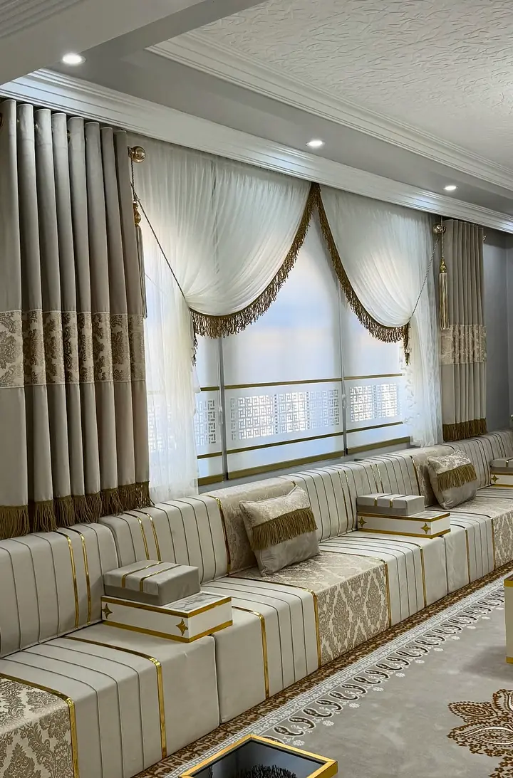 Short ivory damask run below a draped window, with gold-trimmed bases, small fringed pillows and a gold-edged cube table in front