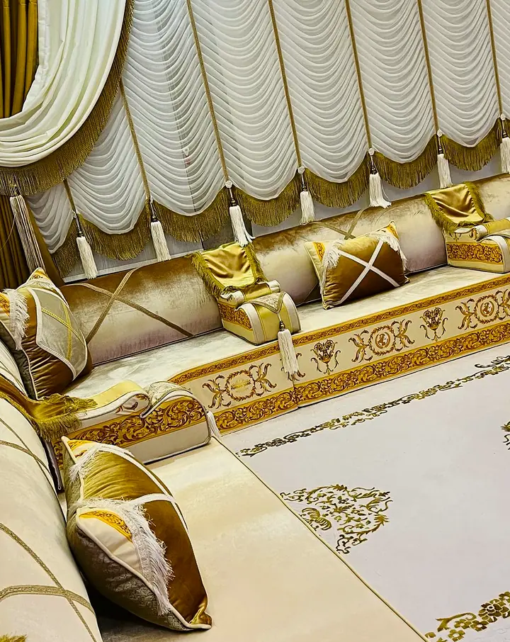Ivory majlis with a gold embroidered band, gold satin pillows with tassels, ruched white curtains and an ivory rug with gold scrolls