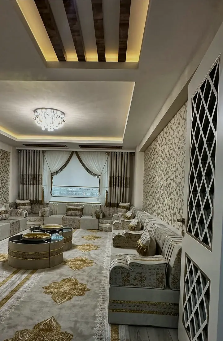 View through an open doorway into an ivory and gold majlis room, with a lattice door panel and drum tables on the rug