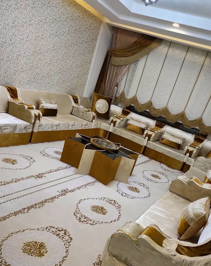 Ivory majlis with gold-brown bases around a star-shaped center table on a cream rug with gold medallions, beside pleated swag curtains