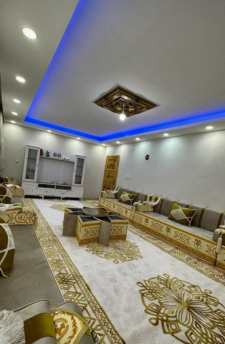 Long hall with two facing runs of gray and olive-gold majlis seating, gold-patterned bands, a star-shaped table and a cream and gold rug