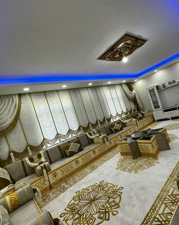 Long living room with a majlis in pale gray and olive-gold under festoon curtains, a star table and a cream rug with gold medallions