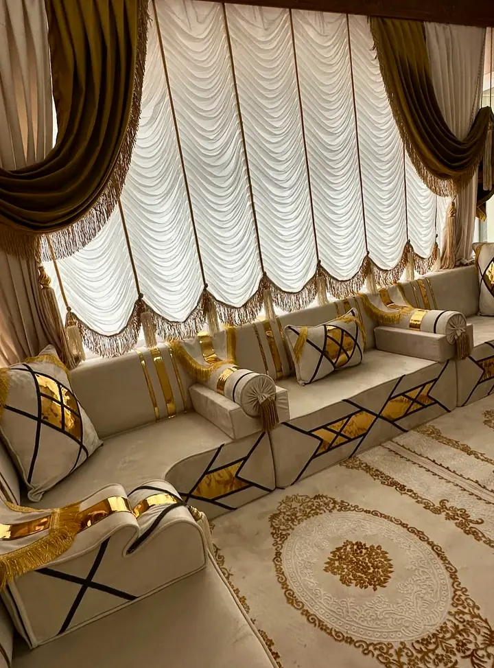 Ivory velvet majlis corner below a festoon blind, with black-line and gold mirror bases, tasseled bolsters and a cream medallion rug
