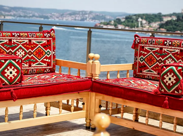 Close view of a wooden bench corner with a red kilim seat cushion, backrest pillows and turned spindle rails