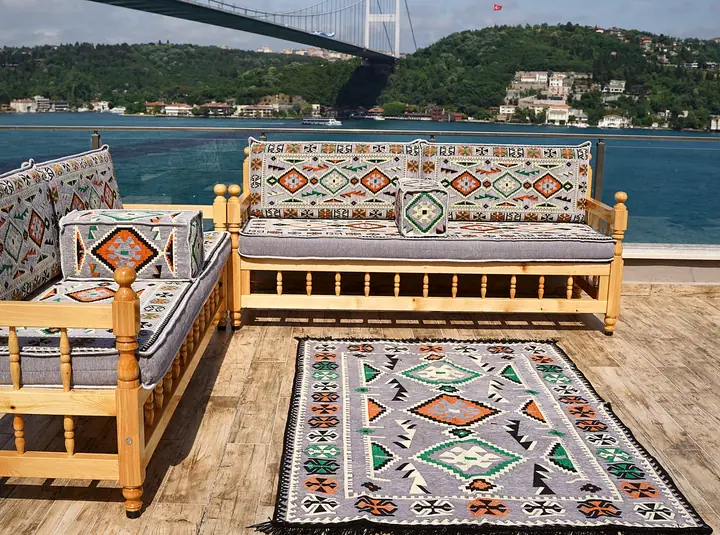 Two wooden benches meeting at a corner in an L, with gray kilim cushions and a patterned rug laid in front