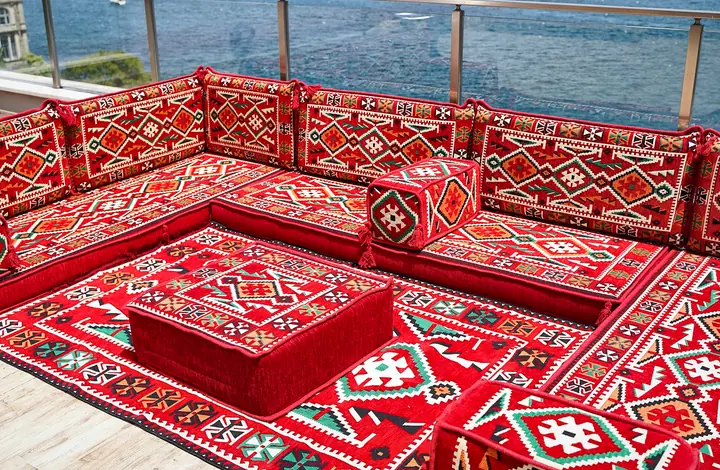 Angled view of red kilim floor seating on three walls with an ottoman standing on the matching rug