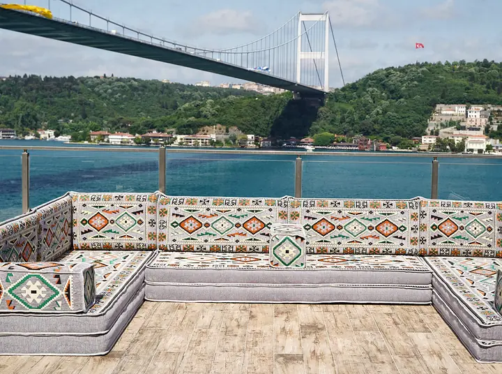 Light gray kilim floor seating on three sides with orange and green accents, set out on a bare wooden terrace above the water