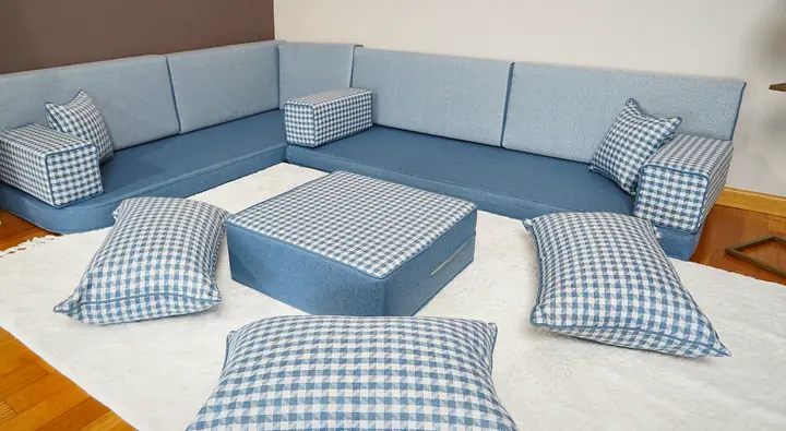 Light blue linen L-shaped corner floor sofa with gingham-check armrests, a gingham ottoman and floor pillows on a white rug