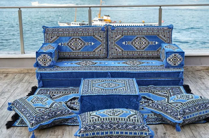 Light blue medallion-pattern single floor sofa with a matching ottoman, four floor pillows and a rug on a waterfront terrace