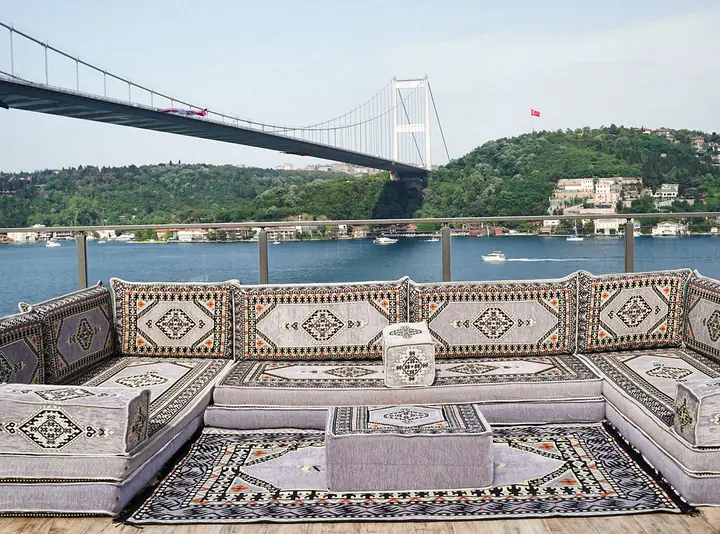 Light gray U-shaped floor sofa in a medallion kilim weave with a matching pouf on a kilim rug, on a terrace below a suspension bridge
