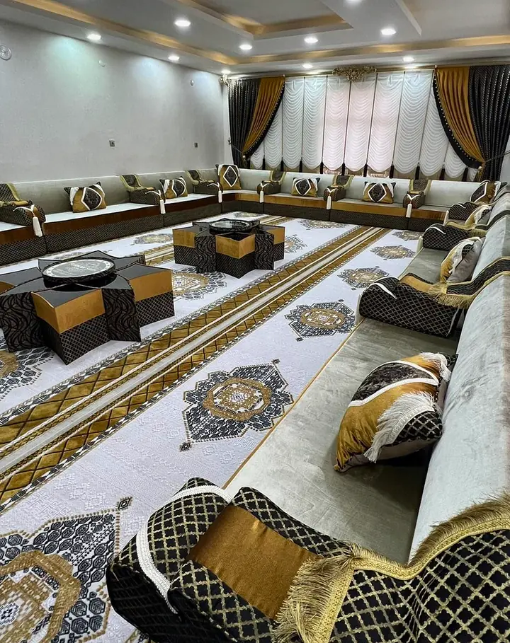Spacious hall with a light gray velvet majlis, black and gold lattice armrests, white swag curtains and a white carpet with gold medallions