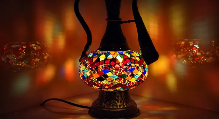 Lit ewer-shaped mosaic lamp with red and amber glass pieces glowing on a dark, reflective tabletop