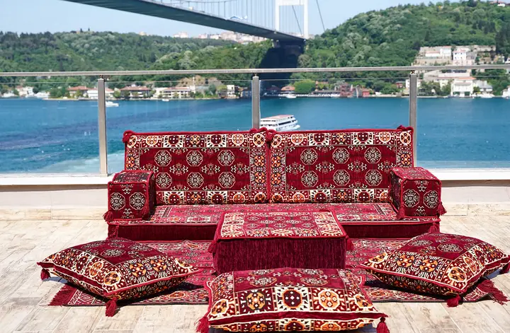 Maroon single floor couch patterned with rows of rosette medallions, set with an ottoman and four floor pillows by the water