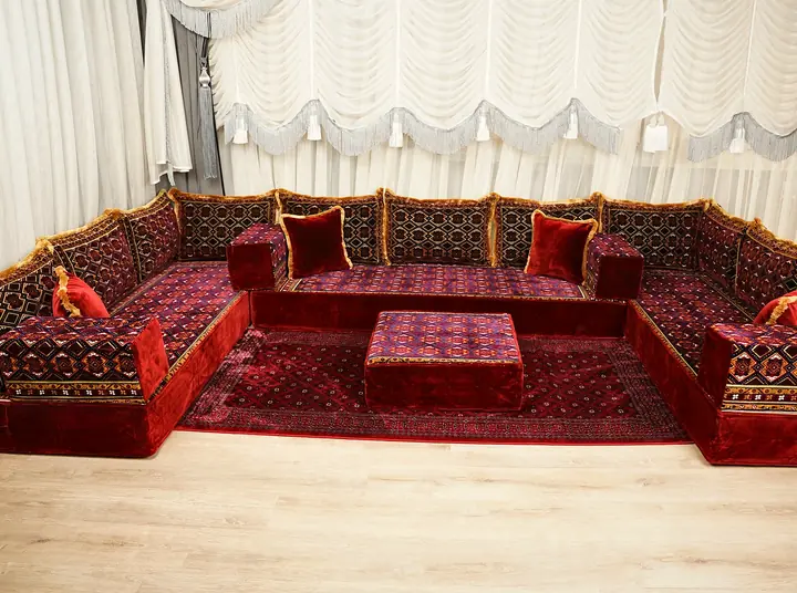 U-shaped maroon velvet floor couch with gold-fringed patterned backrests, red velvet pillows and an ottoman on a red rug