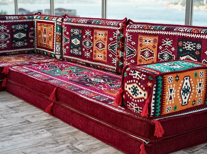 Angled view of an L-shaped red kilim set with a tasseled armrest bolster resting on a gray wood floor