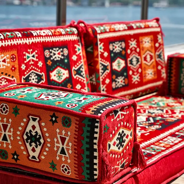 Close view of a red kilim armrest bolster and back cushions, showing orange and green motifs in the weave