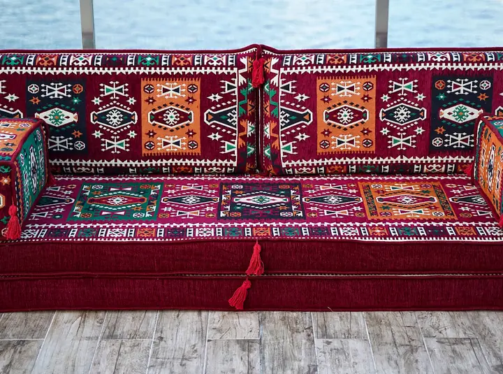 Thick single red kilim floor sofa seen straight on, with teal armrest bolsters and patterned back cushions