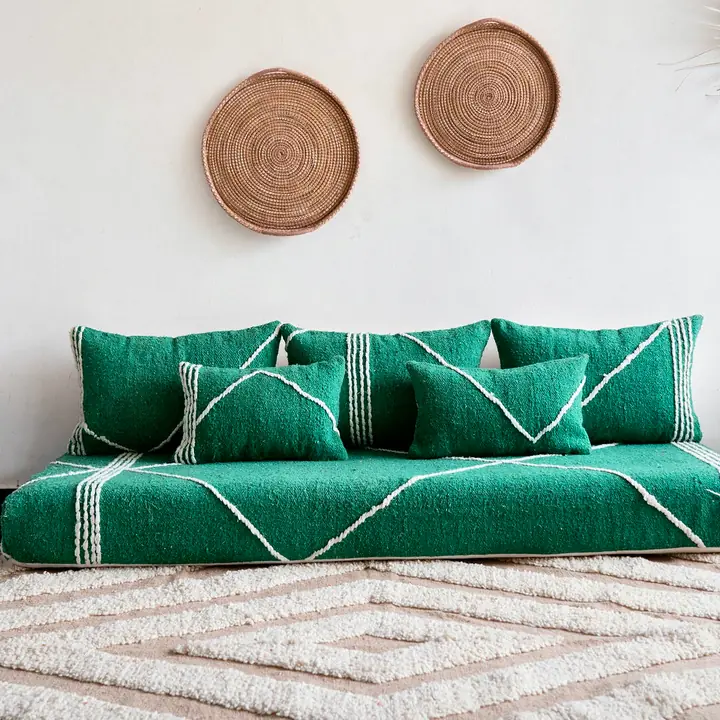 Green cotton floor couch with white stitched lines and matching back pillows on a cream shag rug, two woven baskets on the wall above