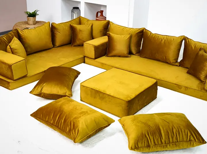 Mustard gold velvet L-shaped floor sofa with a square ottoman and three floor pillows on a white rug in a bright room