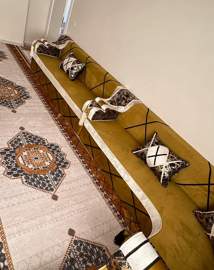 Mustard-gold velvet majlis along a white wall with black diamond lines, gold-and-black pillows and a cream medallion rug