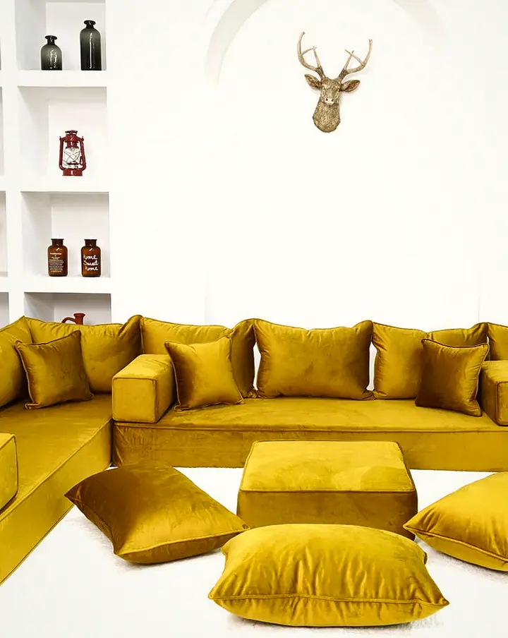 Thick mustard gold velvet U-shaped floor sofa with an ottoman and floor pillows in a bright white room with shelving