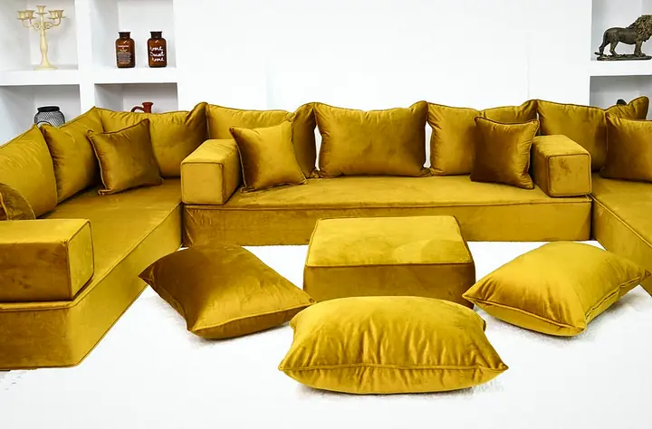 Thick mustard gold velvet U-shaped floor sofa with an ottoman and floor pillows in a bright white room with shelving