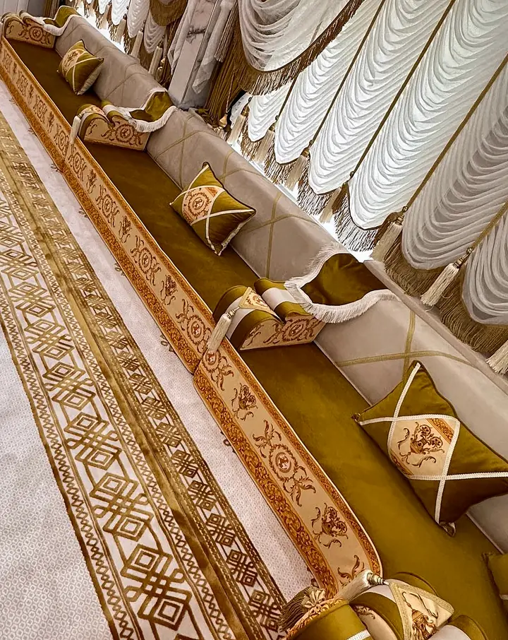 Mustard and ivory majlis along a long wall with gold baroque bands, tasseled armrests, white swag curtains and a gold-bordered rug