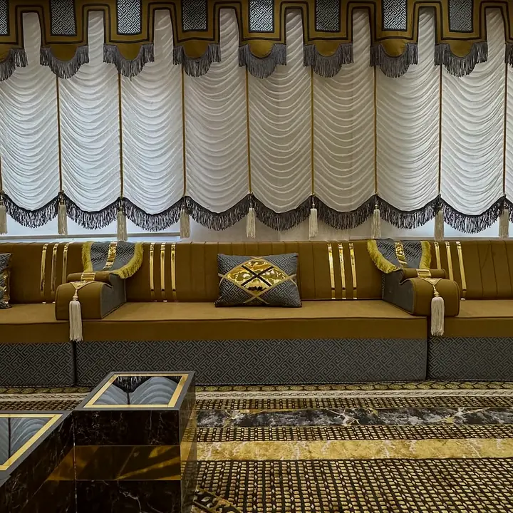 Straight run of mustard seats with Greek-key armrests and patterned pillows below white festoon blinds with gray fringe