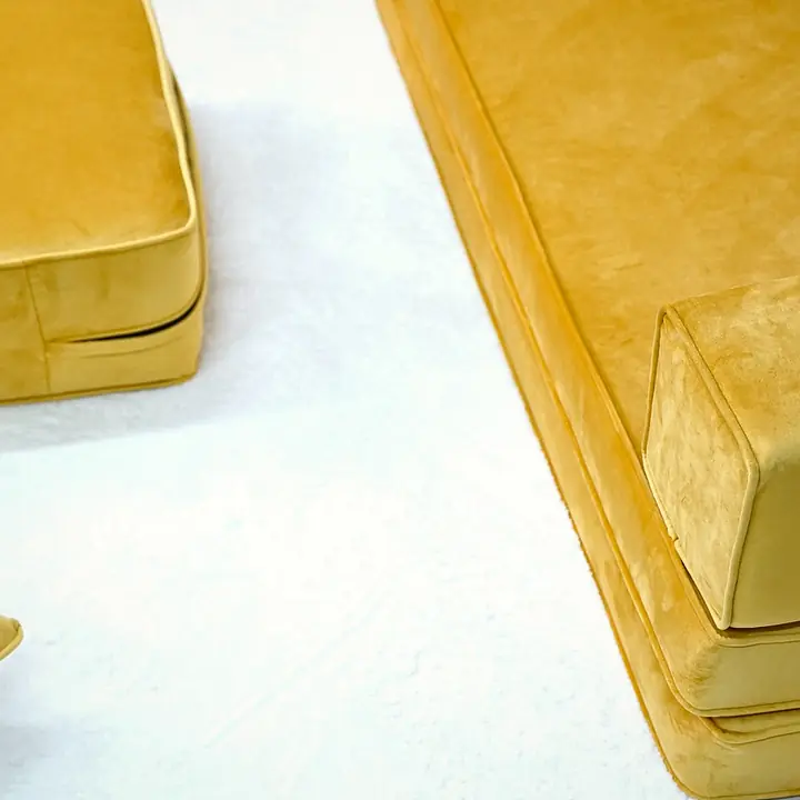 Mustard yellow velvet floor sofa seen from above, showing its layered seat base and a square box armrest beside a stacked pouf