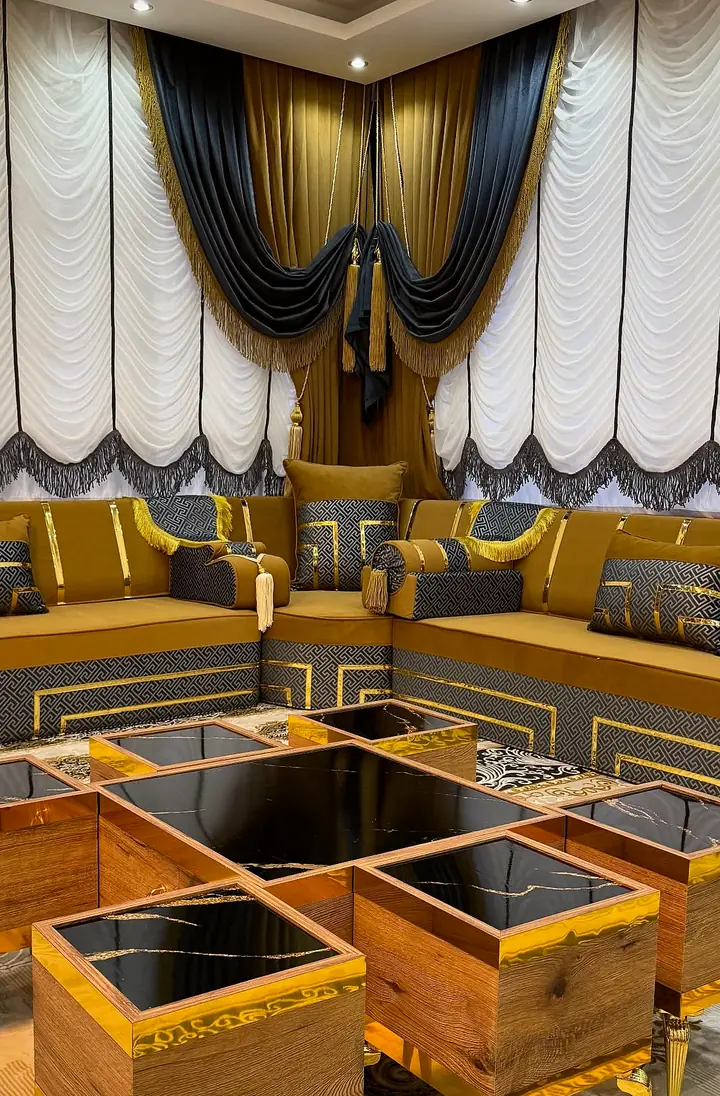 Corner of a mustard velvet majlis with Greek-key armrests and gold fringe, white festoon blinds and charcoal swags above