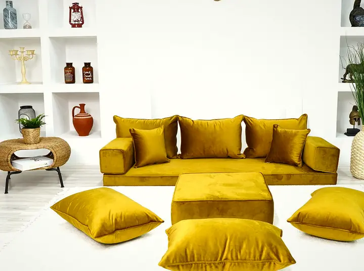 Mustard velvet floor sofa with three back cushions and block armrests, a square ottoman and floor pillows on a white rug in a bright room