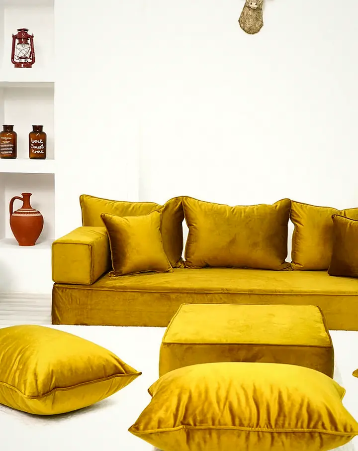 Thick mustard velvet single floor sofa set with a square ottoman and floor pillows on a white rug in a white room with shelving