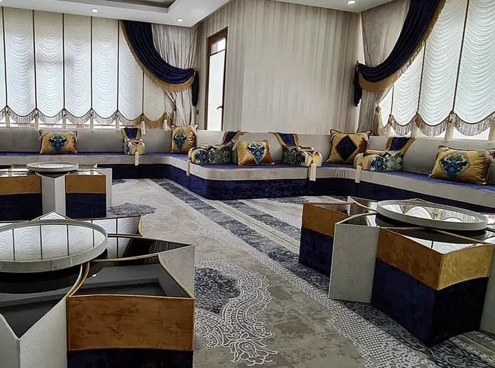 Navy and cream majlis along three walls of a long room with fringed swag curtains, orange-panel star-shaped tables and a patterned rug