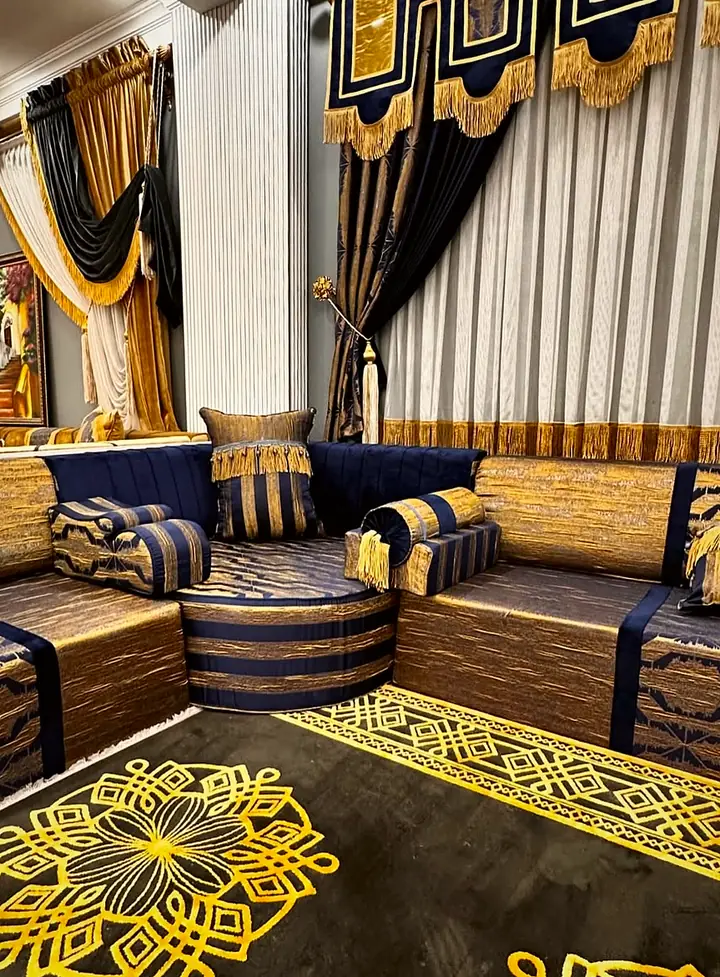 Inside corner of a navy and gold brocade majlis filled by a round pouf, with striped bolsters and a black rug with gold medallions