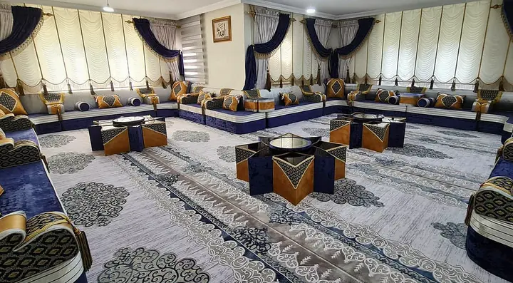 Large room seated wall to wall in navy velvet and silver-grey majlis with burnt-orange cushions and star-shaped tables on a pale carpet