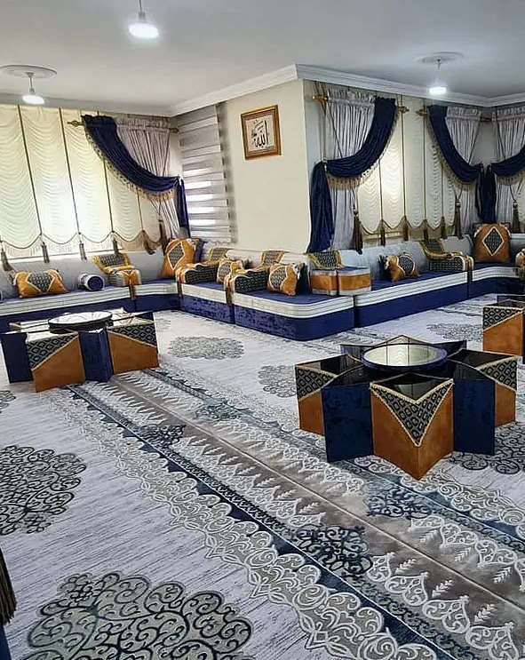 Large room seated wall to wall in navy velvet and silver-grey majlis with burnt-orange cushions and star-shaped tables on a pale carpet