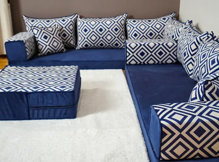 Navy velvet corner floor couch with blue-and-white diamond backrests and a matching ottoman on a white rug, cream curtains at the window