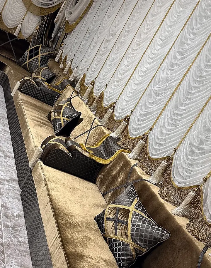 Olive-bronze velvet majlis along a wall with gray lattice-quilted pillows, gold-trimmed cushions and white swag curtains with tassels