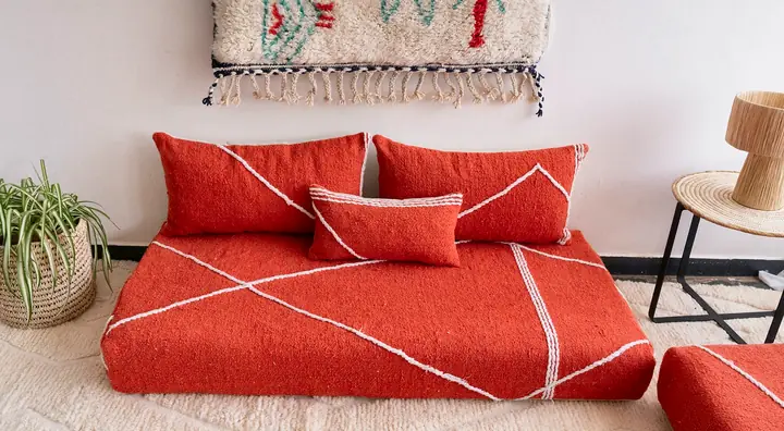 Orange-red Moroccan floor couch with a white line pattern and matching pillows, a woven rug hung on the wall and a rattan lamp