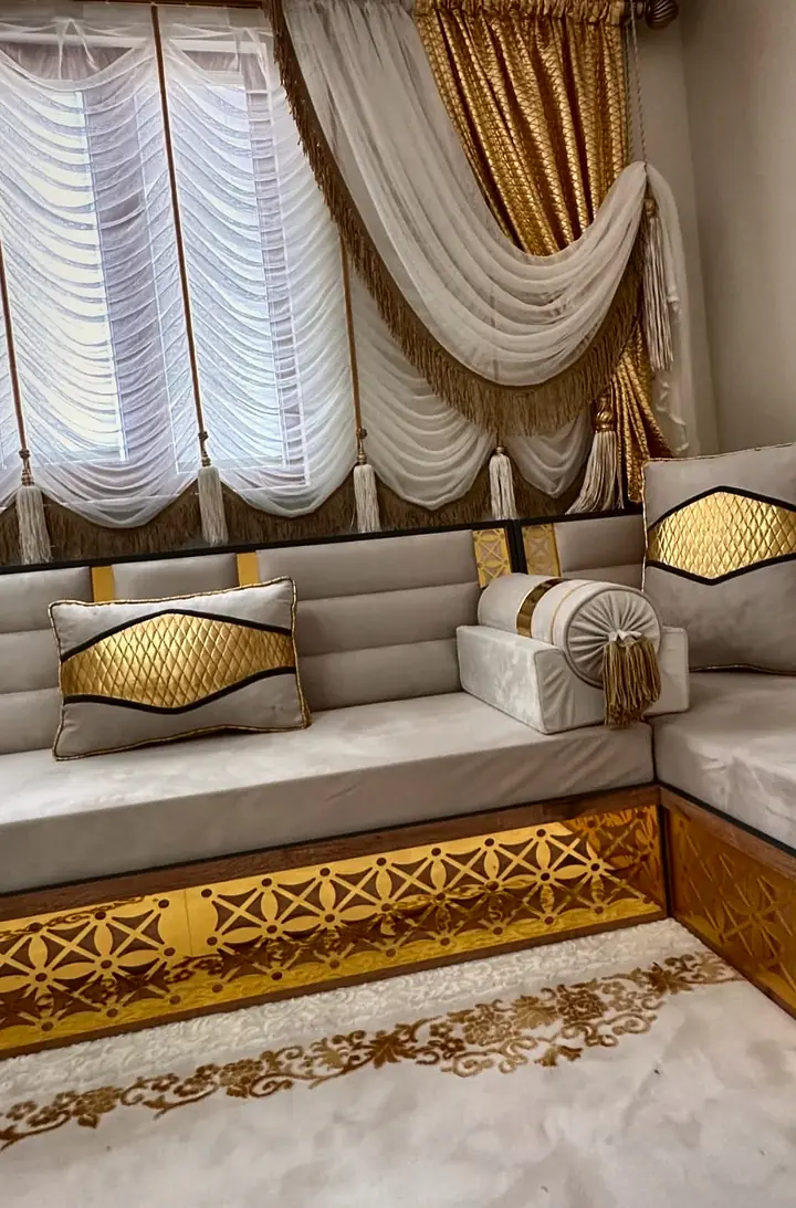 Pale grey raised majlis turning a corner beneath a curtained window, set on a wooden base with gold lattice panels