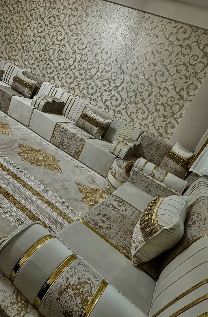 Ivory and taupe seats with gold bands and patterned pillows line a floral wall beside a cream carpet with gold medallions.