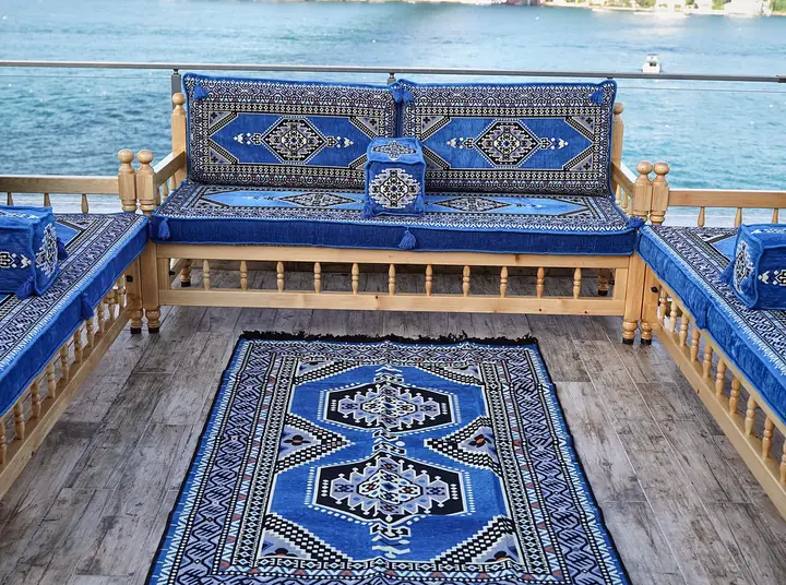 U arrangement of natural pine benches with blue palace-motif cushions around a matching rug, with the sea behind