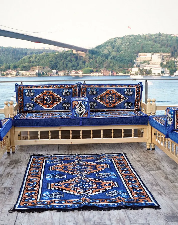 Three pine benches arranged around a patterned rug, with royal blue medallion cushions and a bridge visible through the window